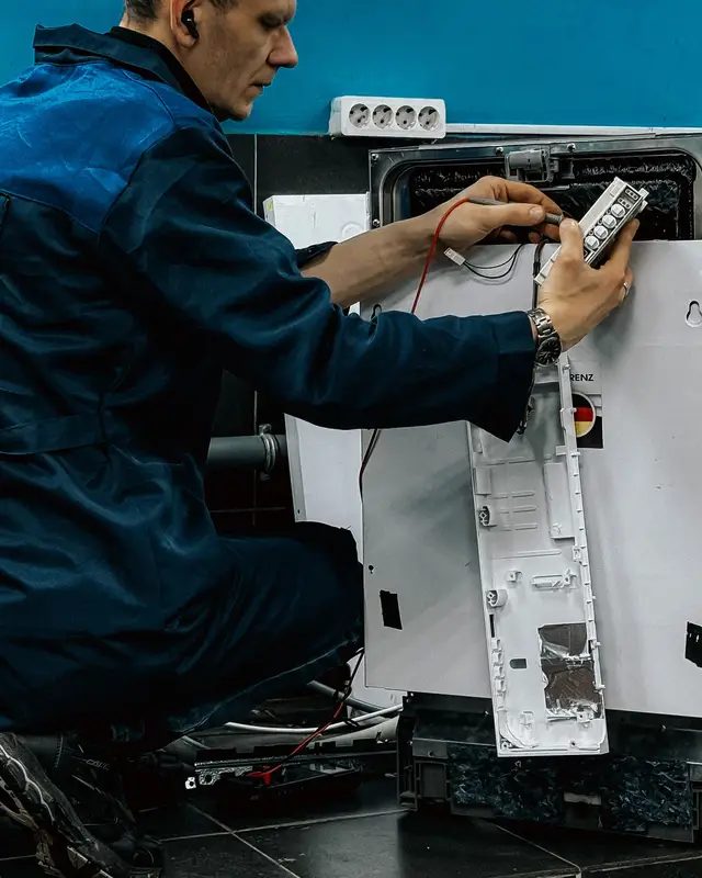 A technician in a blue work uniform kneeling beside a dishwasher with its side panel off, testing its control panel with wires connected
