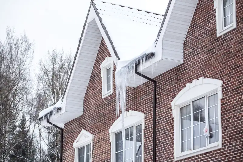 A two-story red brick house in winter with snow on the roof and long icicles hanging from the gutters