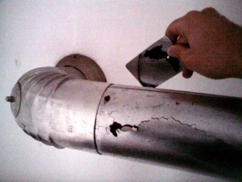 A corroded metal exhaust vent pipe from a gas appliance with holes eaten through it, and a hand holding a piece that came off