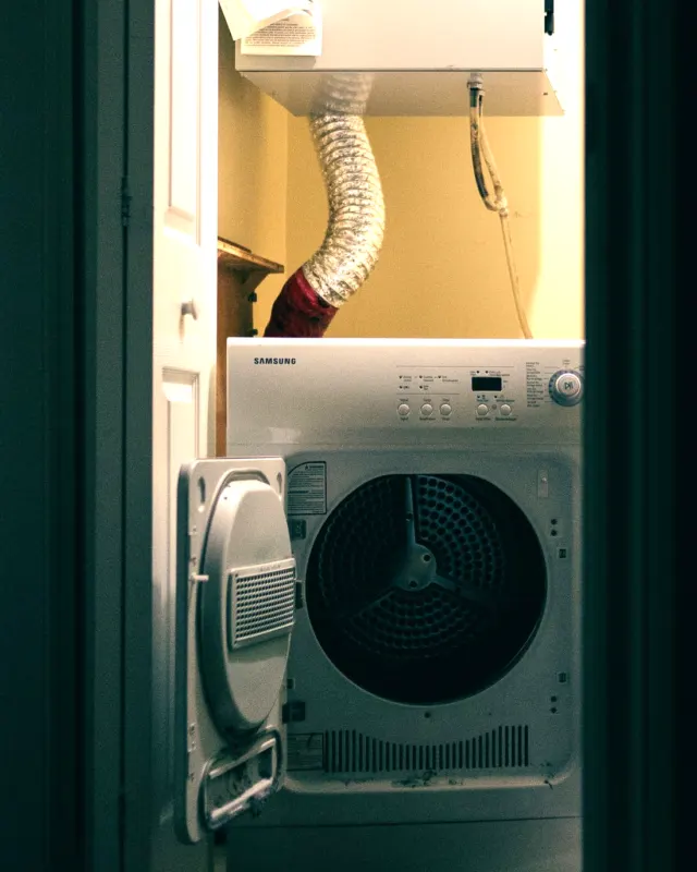 A front-load dryer in a narrow laundry closet with a ribbed foil flexible duct running up the wall behind it