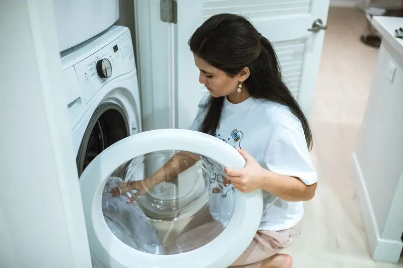 A woman kneeling to load clothes into a front-load washing machine with its round glass door open, in a home laundry closet