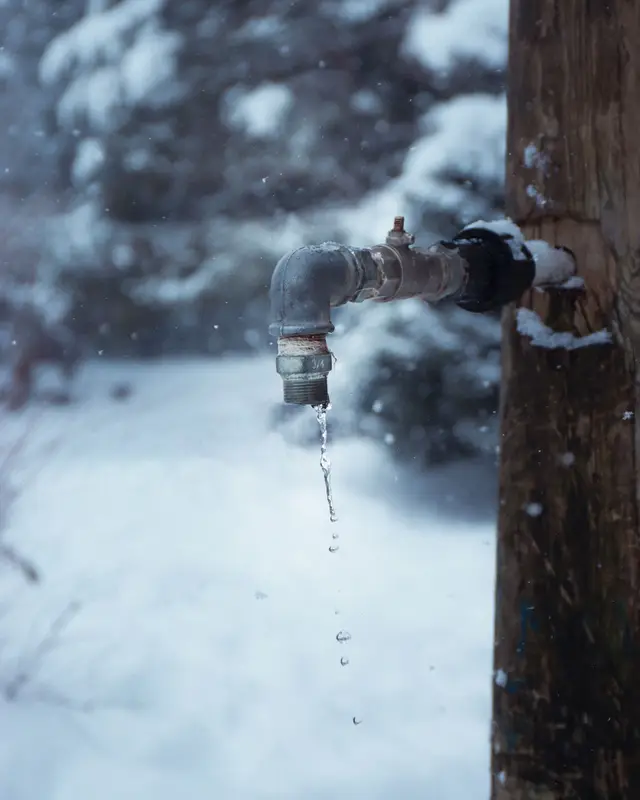 An outdoor metal water faucet on a wooden post in falling snow, with water dripping from the spout