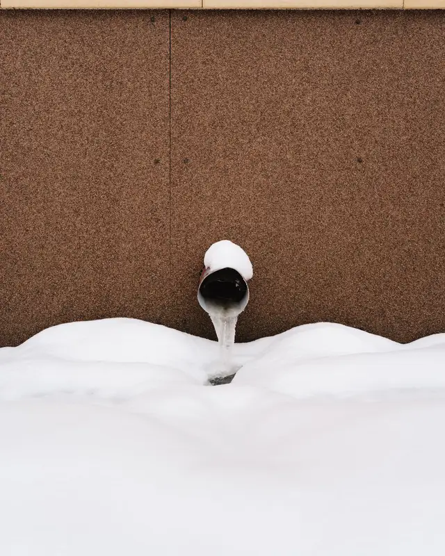 A short pipe coming out of a building wall above deep snow, with ice built up at its mouth