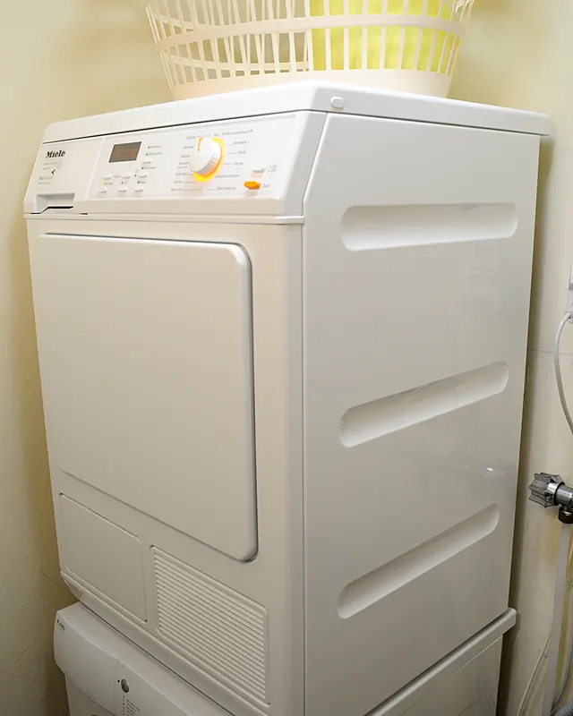 A white heat pump clothes dryer stacked on a washing machine, with a laundry basket on top and no exhaust duct behind it