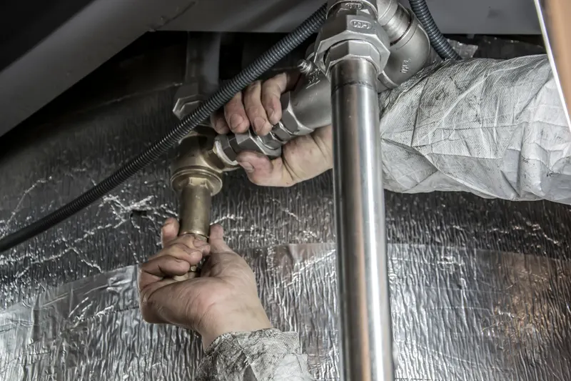 A plumber's hands tightening steel pipe fittings under a cabinet lined with foil insulation