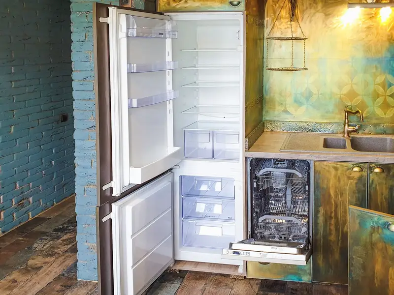 A built-in refrigerator with both doors open and an open built-in dishwasher beside it in a kitchen with a brick wall