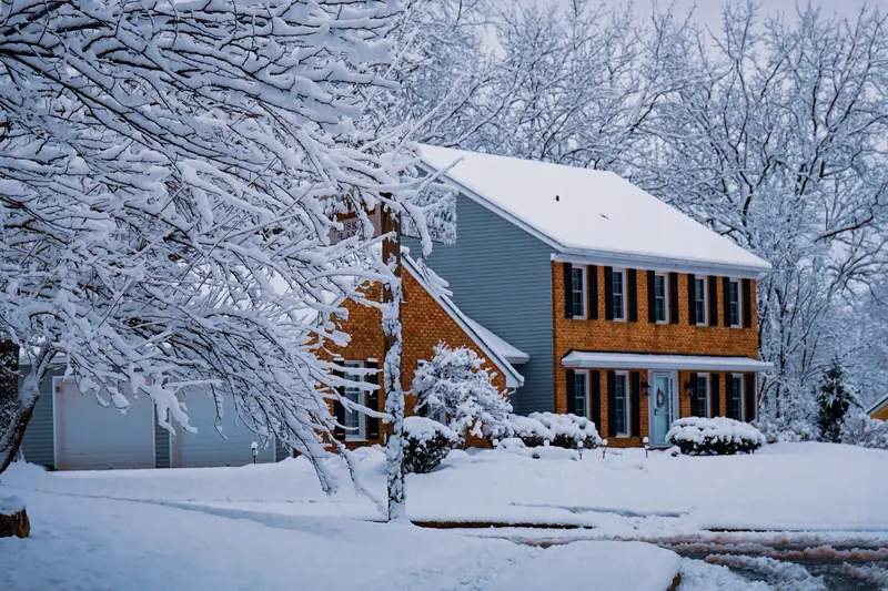 A two-story brick and siding colonial house after a snowfall, with snow-covered trees and a plowed street in front