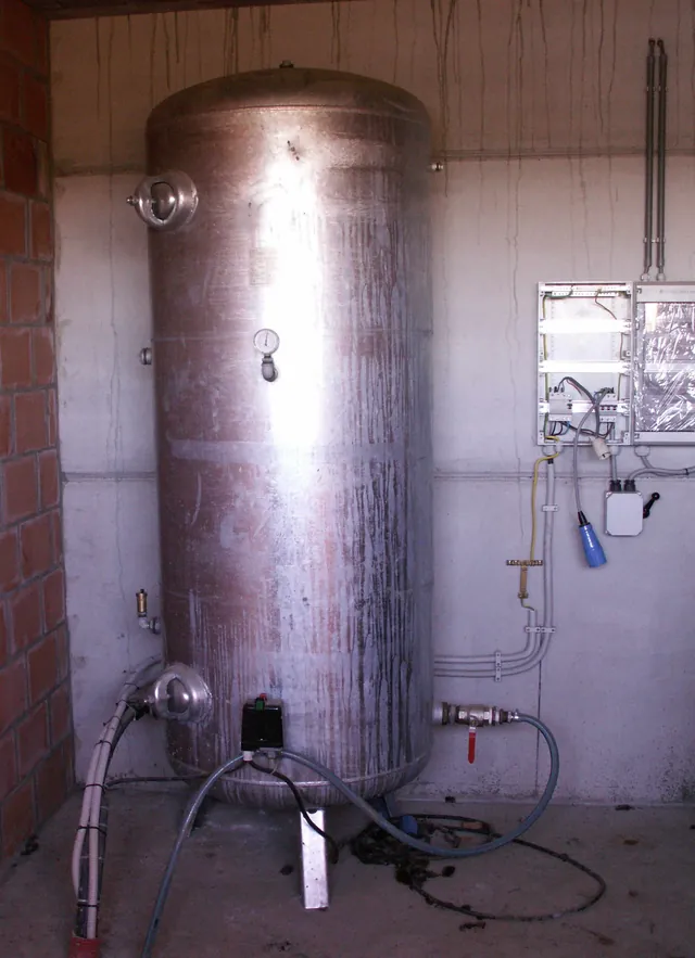 A tall galvanized steel water pressure tank in a utility room, with a pressure gauge on its side and a pressure switch on the pipe at its base