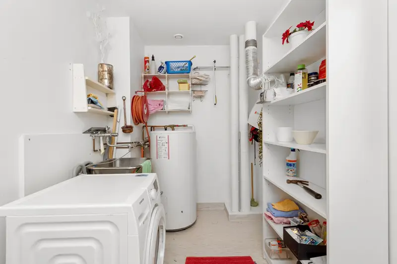 A small utility room with a washing machine, a white tank water heater, a utility sink and storage shelves