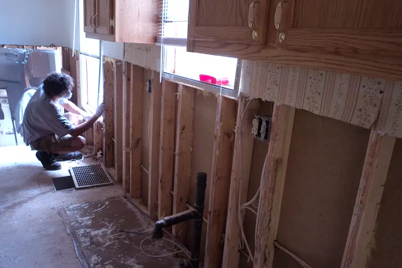 A volunteer in a mask kneels beside wall studs where the lower drywall has been cut away after a flood, with kitchen cabinets above and a floor vent below